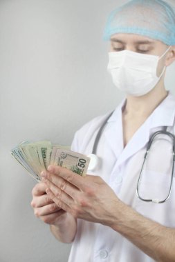 Koruyucu maskeli bir erkek doktor, elinde steteskopla elinde dolar banknotları, sağlık sigortası kavramı, bütçe, tedavi masrafları, organ bağışı hayranı.
