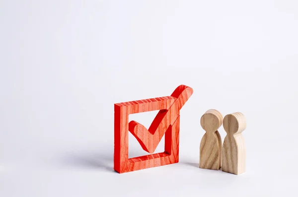 Twee houten menselijke figuren staan samen naast een rood vinkje in het vak. Het concept van verkiezingen en sociale technologieën. Vrijwilligers, partijen, kandidaten, Kieskring kiezers. Rechten van de mens. — Stockfoto