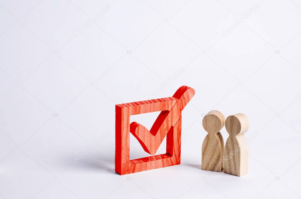 Two wooden human figures stand together next to a Red tick in the box. The concept of elections and social technologies. Volunteers, parties, candidates, constituency electorates. Human rights.