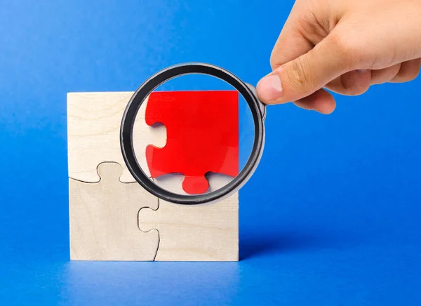 Una lupa mira un rompecabezas de madera roja sobre un fondo azul. Opinión individual. Destacan entre la multitud. Singularidad. Visión divergente. Conceptos diferentes a los de otras personas . —  Fotos de Stock