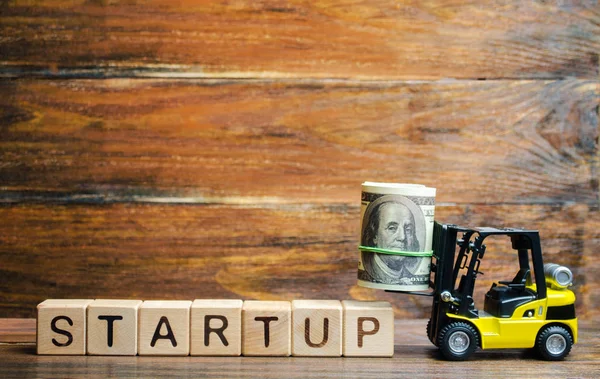 Forklift truck carries a bundle of dollars to inscription Startup. The concept of raising funds for a startup. Crowdfunding. Raising funds and resources to complete a commercial or intangible project.