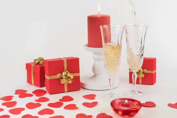 Due bicchieri di champagne su sfondo bianco. Decorato con candele accese, cuori rossi, scatole rosse con regali. Lo champagne viene versato in un bicchiere. Il giorno di S. Valentino. Biglietto per anniversario. Copyspace — Foto Stock