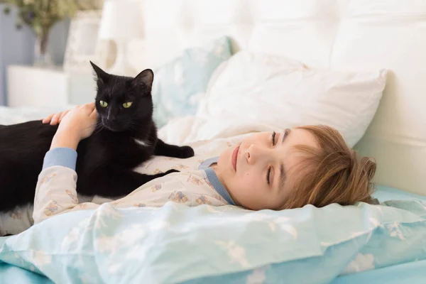 Beyaz çocuk parlak bir odada bir yatakta uyur. Siyah kedi bebeklerle sarılıp uyuyor.