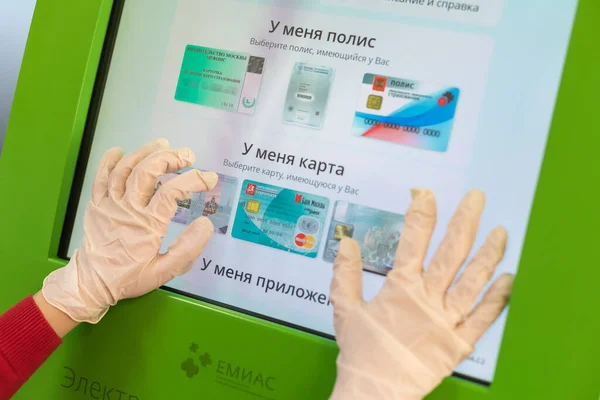 26.03.2020 Rusya, Moskova. Hastanın elleri EMIAS terminalinde koruyucu eldivenlerle doktorla kayıt yapıyor. Sağlık sigortası poliçesi seçeneğiyle birlikte terminal skorbordu.