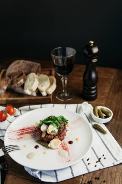 Biftek tartare bıldırcın yumurtası, turşu ve roka, ekmek ve bir kadeh kırmızı şarap koyu ahşap bir masa üzerinde