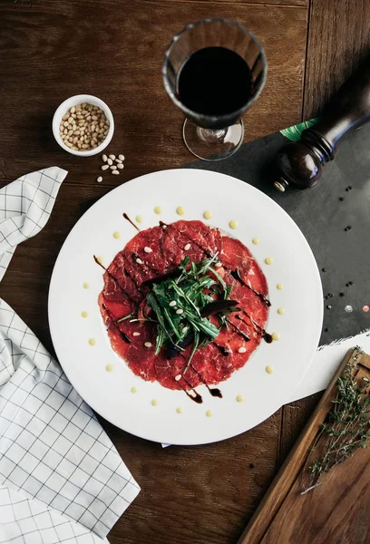 Beef Carpaccio Rukolą Białe Płytki Kieliszkiem Czerwonego Wina Orzeszkami Pinii — Zdjęcie stockowe