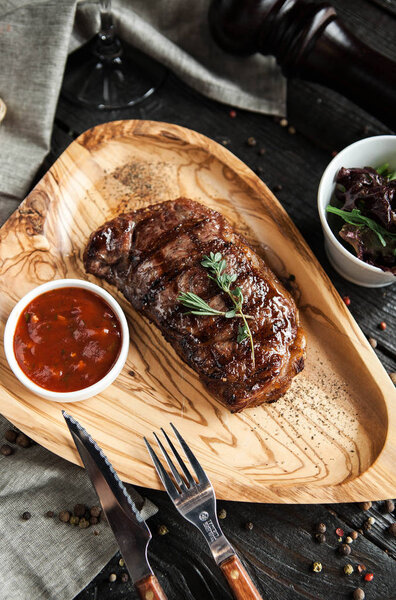 Beautiful serving of meat steak on a wooden plate with salad, tomato sauce and a glass of red wine on a wooden dark background