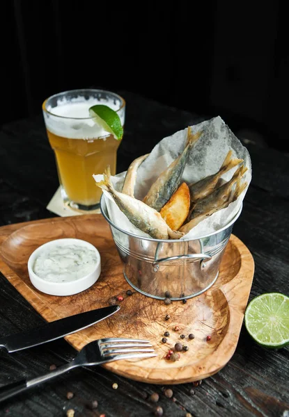 snack to beer - fried small fish with potatoes and sauce on a wooden plate with a glass of light beer