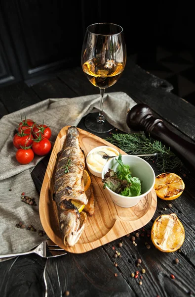 美丽的烤鱼与柠檬在一个木盘子与一杯白色的葡萄酒在一个黑暗的木质背景 — 图库照片