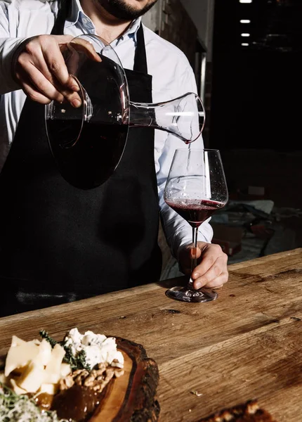 在一个黑色的围裙和一个轻衬衫的品酒师倒入红酒从一个滗水器成玻璃 — 图库照片
