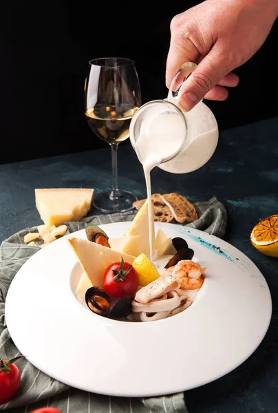Man's hand pours a cheese soup with seafood, mussels, shrimp, squid with lemon and croutons in a beautiful serving with a glass of white wine — Stock Image Man Hand Pours Cheese Soup Seafood Mussels Shrimp Squid Lemon Stock Photo
