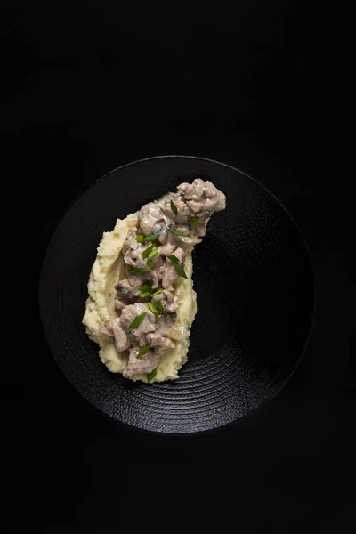 Μεσημεριανό μοσχάρι στρογκανόφ με πουρέ πατάτας και πράσινα κρεμμύδια σε μαύρο πιάτο και σε μαύρο φόντο Εικόνα Αρχείου