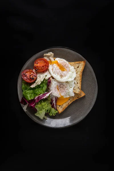 Πρωινό με δύο φρυγανιές λευκού ψωμιού, φύλλα σαλάτας, ντοματίνια και δύο αυγά ποσέ σε γκρι πιάτο σε μαύρο φόντο Royalty Free Εικόνες Αρχείου