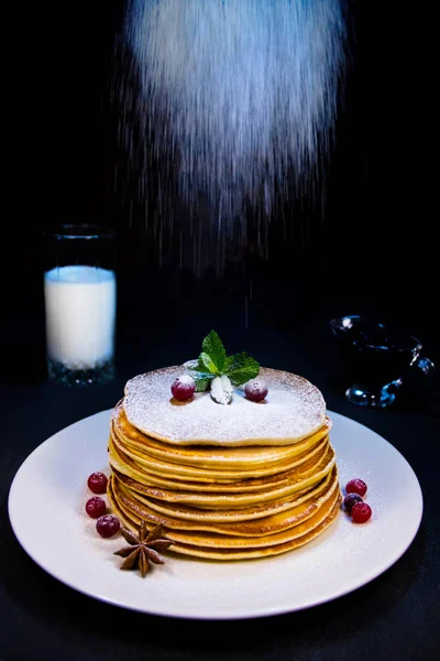 Pancakes on a white plate with cranberry puddings and cinnamon sprinkled with sugar powder along with blueberry jam and a glass of milk — Stock Photo, Image