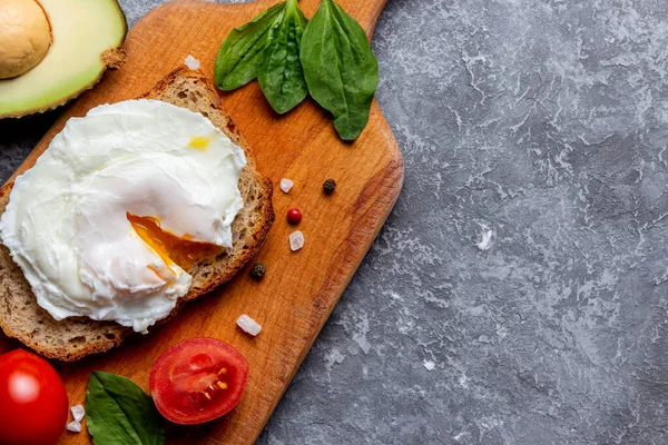 Poached egg on bread with seeds, cherry tomato, spinach, avocado, salt and spices on a wooden tray on stone background