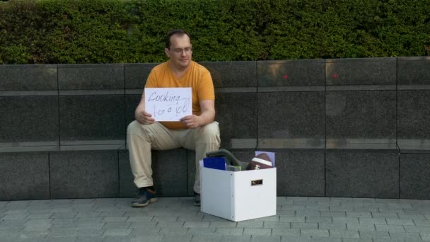 El desempleado en la calle con corton con inscripciones necesita trabajo. El hombre despedido se sienta en una calle de hormigón con una caja con pertenencias personales . — Vídeos de Stock