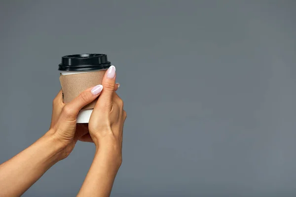 Тема для завтрака и кофе: женская рука держит белый бумажный кофейный стаканчик с черным пластиковым чехлом на сером фоне в студии, рекламирует кофе. Копирование пространства, серый фон, изолировать — стоковое фото
