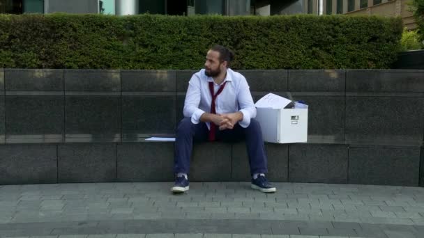 Werkloze lachende man op straat. Ontslagen man zit op een betonnen straat met een doos met persoonlijke bezittingen. — Stockvideo