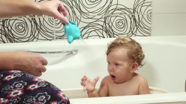 O garoto toma banho no banheiro com sua mãe e brinca com brinquedos. Um bebê de olhos azuis com cabelos loiros banha-se com brinquedos sob a supervisão de sua mãe. Lavar e banhar crianças, Higiene e — Vídeo de Stock