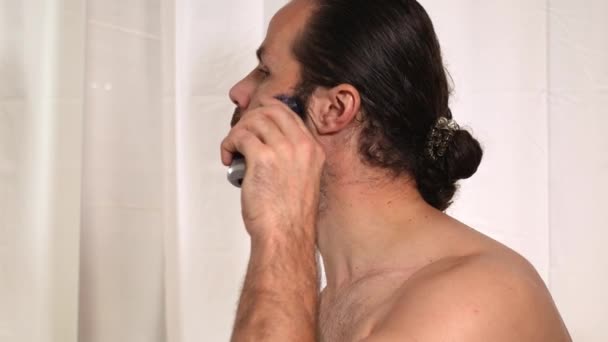 Красивий молодий чоловік з довгим волоссям голить бороду з тримером у ванній. Крупним планом . — стокове відео