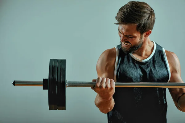 Retrato de un joven musculoso súper en forma haciendo ejercicio en el gimnasio con barra de pesas sobre fondo gris, copyspace . —  Fotos de Stock