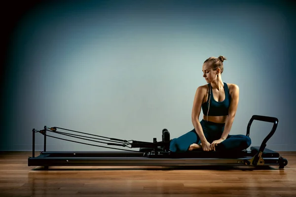 Chica joven haciendo ejercicios de pilates con una cama reformadora. Hermoso entrenador de fitness delgado sobre fondo gris reformador, bajo llave, luz de arte. Concepto Fitness — Foto de Stock