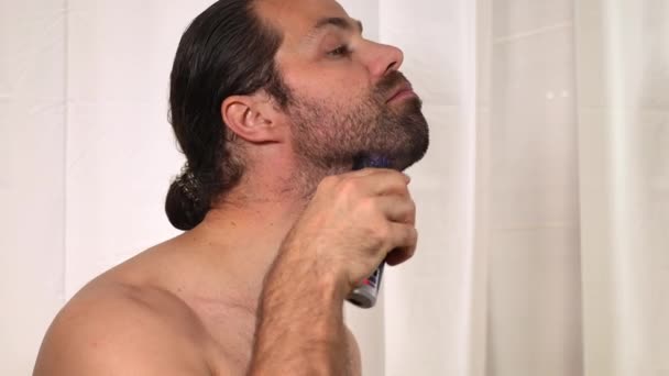 Jóképű, hosszú hajú fiatalember borotválja a szakállát egy nyíróval a fürdőszobában. Közelkép. — Stock videók