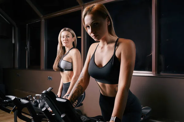 Duas mulheres desportivas atraentes andar de bicicleta de exercício durante o treinamento de ciclismo no ginásio — Fotografia de Stock