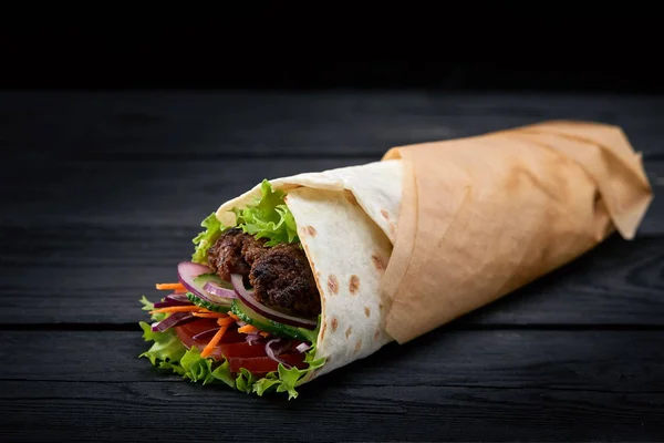 Shawarma rolled in lavash, moist grilled meat with onion, herbs and vegetables on wooden black background. — Stock Photo, Image