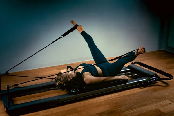 Chica joven haciendo ejercicios de pilates con una cama reformadora. Hermoso entrenador de fitness delgado sobre fondo gris reformador, bajo llave, luz de arte. Concepto Fitness — Foto de Stock