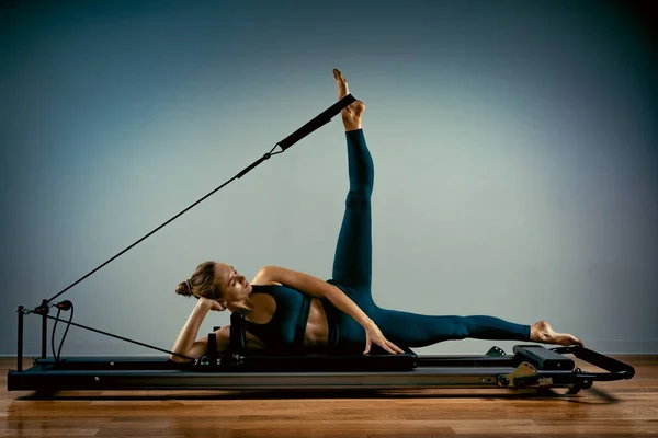 Chica joven haciendo ejercicios de pilates con una cama reformadora. Hermoso entrenador de fitness delgado sobre fondo gris reformador, bajo llave, luz de arte. Concepto Fitness — Foto de Stock