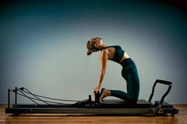 Chica joven haciendo ejercicios de pilates con una cama reformadora. Hermoso entrenador de fitness delgado sobre fondo gris reformador, bajo llave, luz de arte. Concepto Fitness —  Fotos de Stock