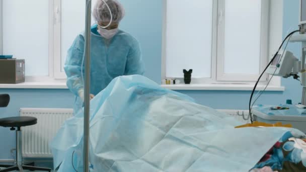 Paciente feminina deitada sob anestesia amarrada na mesa cirúrgica, cirurgiã e assistente preparam o paciente e providenciam esterilidade na sala de cirurgia. — Vídeo de Stock