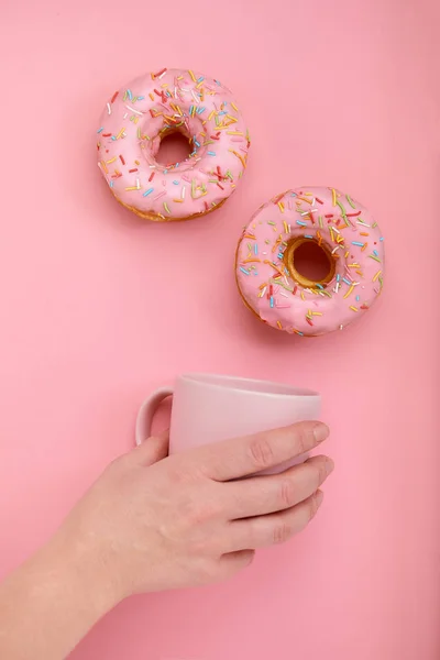 Süße Donuts auf rosa Hintergrund weibliche Hand hält einen rosa Becher mit Kaffee. Farbspiel, Farbkonzept, Einfarbstilisierung. — Stockfoto