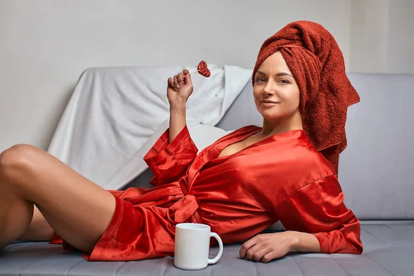 Model in a red bathrobe and a red towel on the sofa with a coffee mug and a lollipop. The concept of fashion, the game of color, minimalism. Rest from work, rest time. — 스톡 사진