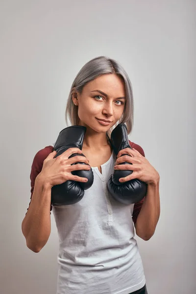 Young beautiful sexy woman boxer posing with black boxing gloves in her hands on a gray background. Copy space, gray background, goal achievement. — Stock Photo, Image