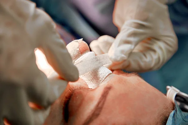 Un chirurgien avec des gants lui opère le nez. Rhinoplastie, opération de gros plan. Lumière bleue, travail d'équipe, santé, beauté . — Photo