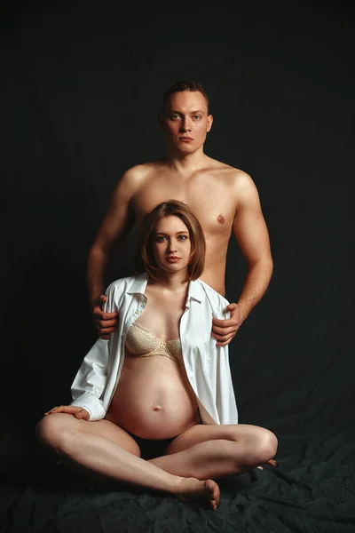 Una pareja embarazada posando ante la cámara sobre un fondo oscuro, discreto. Concepto de embarazo de moda. Copiar espacio, fondo negro . — Foto de Stock
