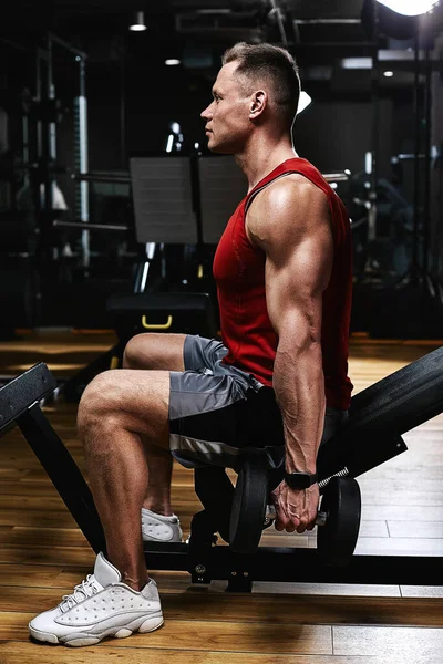 Jonge sporter fitness model in de sportschool schudt zijn biceps met halters. Sportmotivatie, lage toonaard, hoog contrast. Het concept van een gezonde levensstijl, levensbeweging, kopieerruimte. — Stockfoto