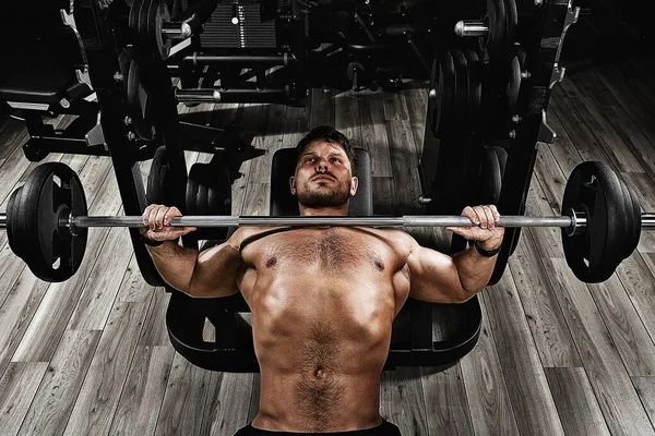 Un joven musculoso levantando un banco de pesas en el gimnasio. Hermoso cuerpo, logro de objetivos, el deporte como una forma de vida . —  Fotos de Stock