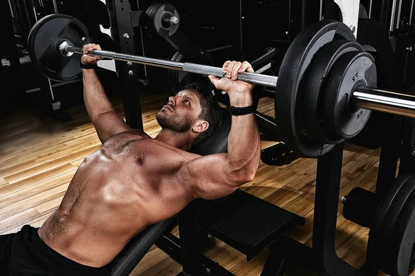 Jonge gespierde man tilt een halterbank op in de sportschool. Sport, beweging, leven. Het concept van een gezonde levensstijl, regelmatige training, ontwikkeling van de borstspieren — Stockfoto