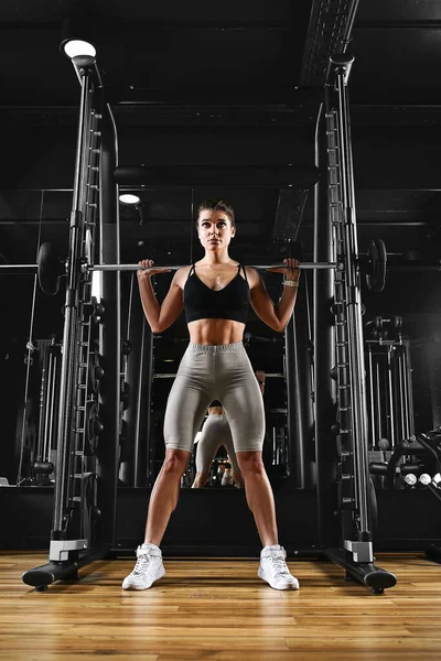 一个女孩正在体育馆用杠铃训练她的腿。运动生活,健身,健身动机.黑暗背景，复制空间 — 图库照片