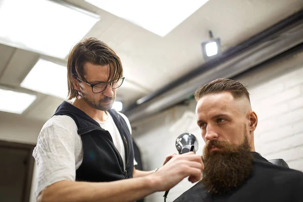 Brutaler Typ im modernen Friseurladen. Friseur frisiert einen Mann mit langem Bart. Friseurmeister frisiert mit Schere und Kamm — Stockfoto