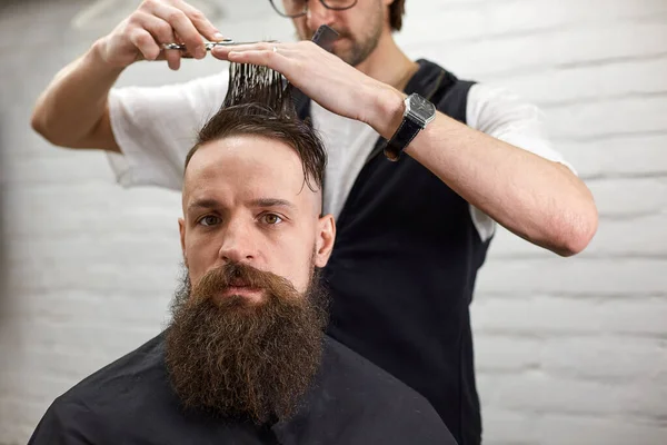 Master cuts hair and beard of men in the barbershop, hairdresser makes hairstyle for a young man