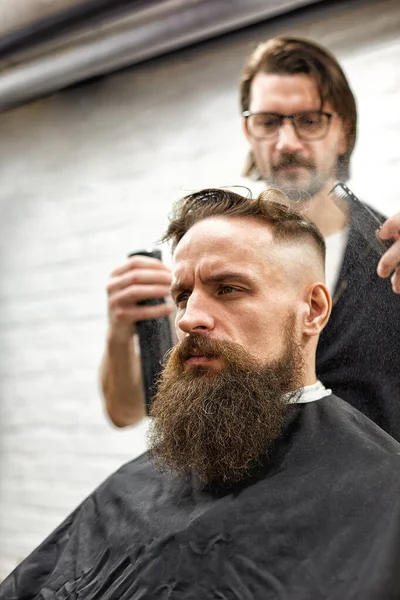 Brutaler Typ im modernen Friseurladen. Friseur frisiert einen Mann mit langem Bart. Friseurmeister frisiert mit Schere und Kamm — Stockfoto