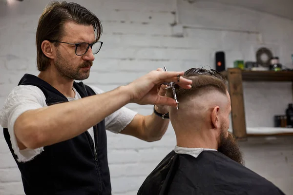 Master cuts hair and beard of men in the barbershop, hairdresser makes hairstyle for a young man