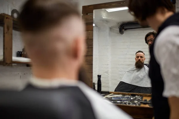 Un tipo brutale nel moderno negozio di barbieri. Parrucchiere rende acconciatura un uomo con una lunga barba. Master parrucchiere fa acconciatura con tagliacapelli — Foto Stock