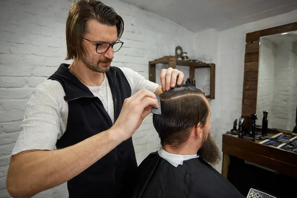 Жестокий парень в современной парикмахерской. Парикмахер делает прическу мужчине с длинной бородой. Мастер парикмахер делает прическу с клиппером для волос — стоковое фото