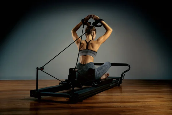 Young girl doing pilates exercises with a reformer bed. Beautiful slim fitness trainer on reformer gray background, low key, art light. Fitness concept — Stock Photo, Image