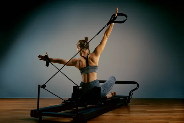 Young girl doing pilates exercises with a reformer bed. Beautiful slim fitness trainer on reformer gray background, low key, art light. Fitness concept — Stock Photo, Image